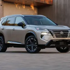 Elegancki, beżowy Nissan Rogue z czarnym dachem i przyciemnianymi felgami stoi przed nowoczesnym budynkiem.