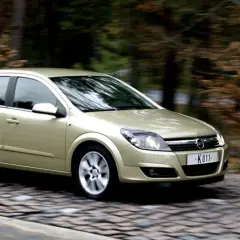 Złoty Opel Astra H 1.7 CDTI w ruchu na brukowanej drodze, otoczony jesiennym lasem.