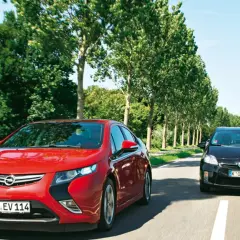 Czerwona Opel Ampera i czarny samochód jadą drogą otoczoną drzewami.