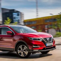 Czerwony Nissan Qashqai 1 jedzie ulicą w mieście. W tle nowoczesne budynki i niebieski znak drogowy.