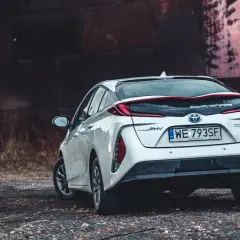 Biały Prius 4 z napędem hybrydowym stoi na tle industrialnego krajobrazu.