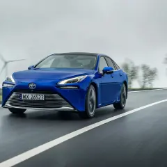 Niebieski sedan z napędem wodorowym jedzie po mokrej drodze, w tle widać wiatraki i drzewa.