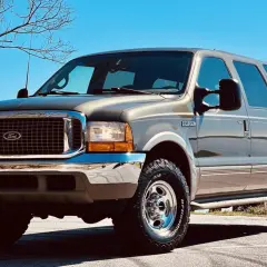 Brązowy Ford Excursion z chromowanymi dodatkami i terenowymi oponami stoi na tle błękitnego nieba i nagich drzew.