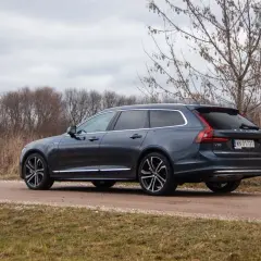 Ciemnoszary Volvo V90 T8 na drodze gruntowej, otoczony suchą trawą i drzewami.