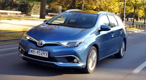 Niebieski Toyota Auris 1.8 VVT-i Hybrid w wersji kombi jedzie po drodze w otoczeniu jesiennych drzew.
