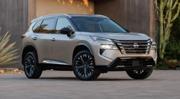 Elegancki, beżowy Nissan Rogue z czarnym dachem i przyciemnianymi felgami stoi przed nowoczesnym budynkiem.