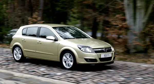 Złoty Opel Astra H 1.7 CDTI w ruchu na brukowanej drodze, otoczony jesiennym lasem.