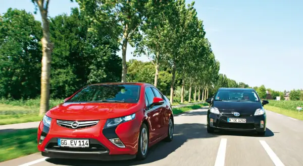 Czerwona Opel Ampera i czarny samochód jadą drogą otoczoną drzewami.