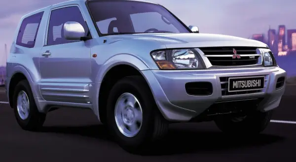 Srebrny Mitsubishi Shogun 3-drzwiowy na drodze o zmierzchu, z miejskim krajobrazem w tle.