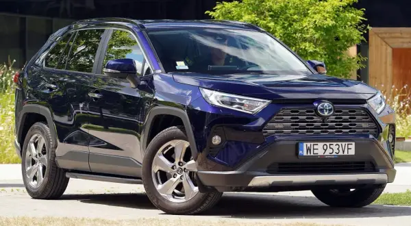 Elegancka, granatowa Toyota RAV4 5 generacji z przyciemnianymi szybami i chromowanymi felgami, zaparkowana na tle zieleni i nowoczesnego budynku.