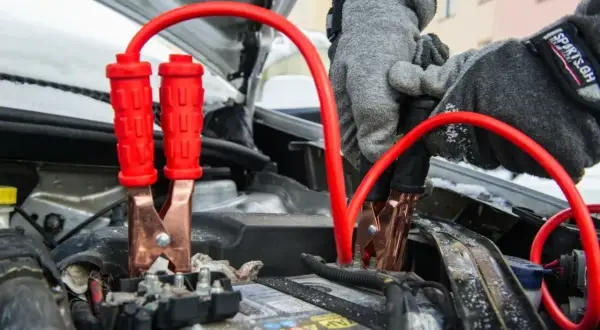 Rękawiczki w szarej zimowej kurtce podłączają kable rozruchowe do akumulatora, pokazując, jak odpalic auto w mroźny dzień.