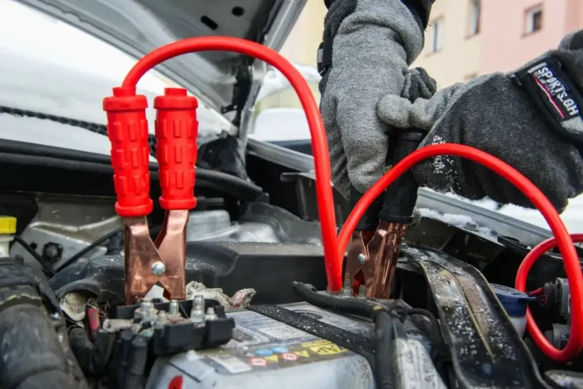 Rękawiczki w szarej zimowej kurtce podłączają kable rozruchowe do akumulatora, pokazując, jak odpalic auto w mroźny dzień.