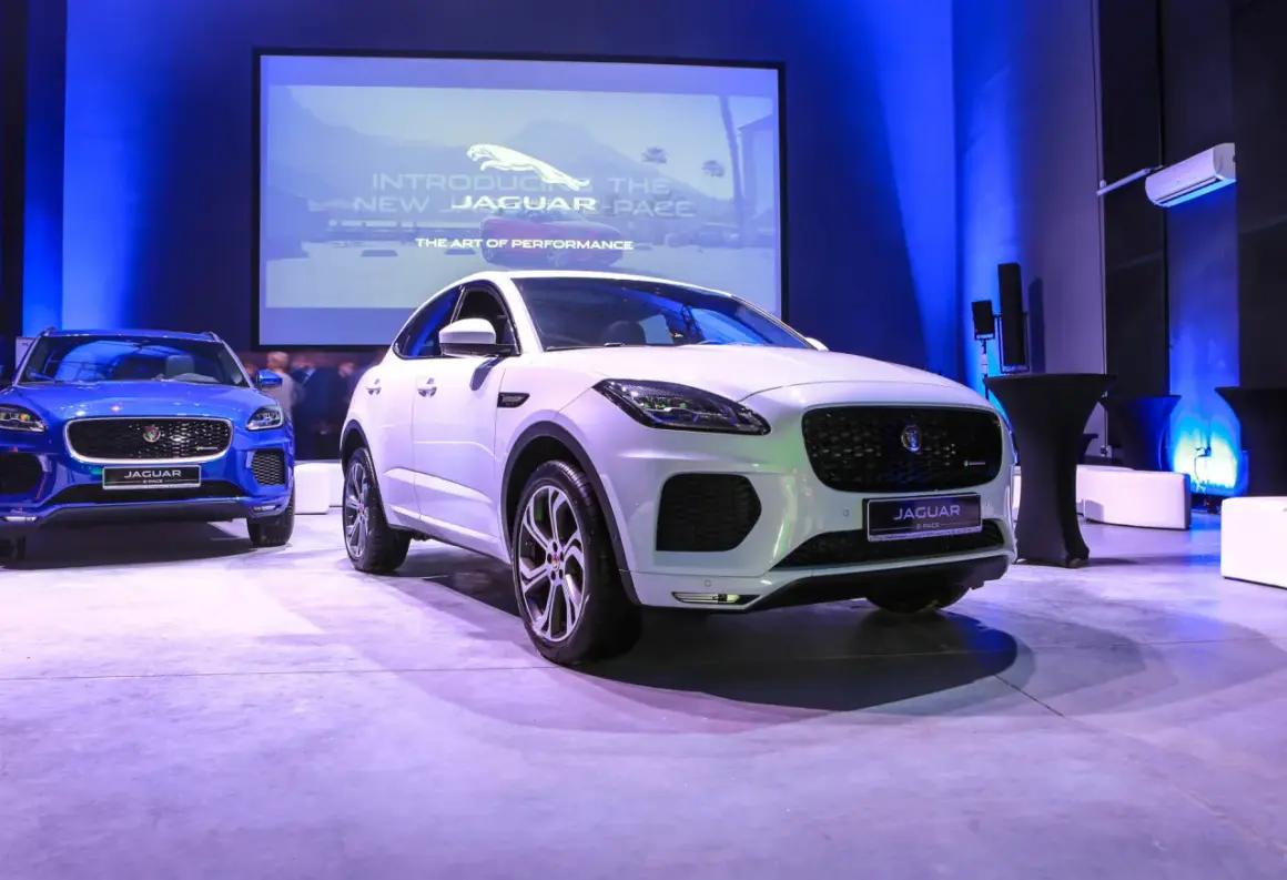 Biały i niebieski Jaguar E-Pace na prezentacji. Na ekranie w tle logo Jaguara i napis "Introducing the new Jaguar E-Pace".