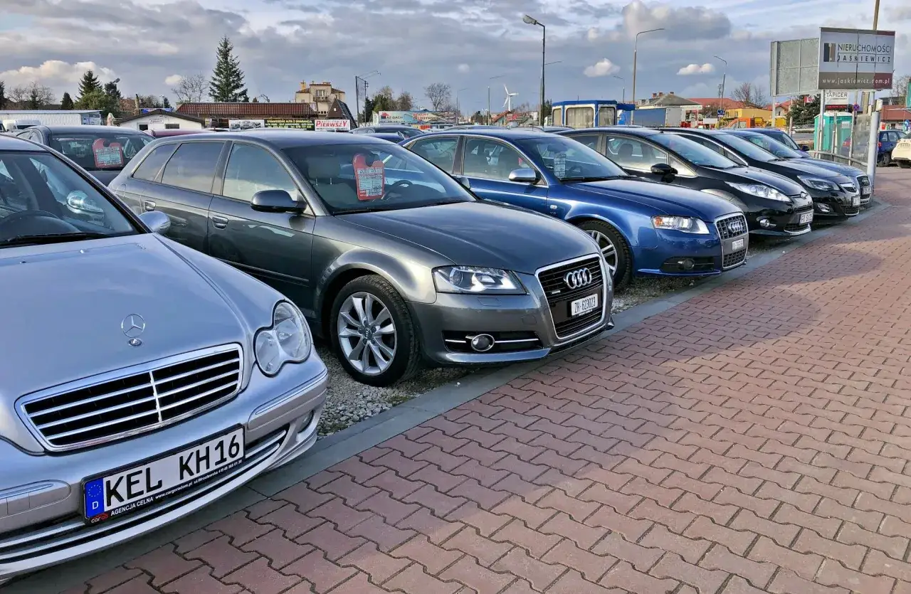Rząd samochodów na placu. Widać srebrnego Mercedesa, szare Audi, niebieskie Audi i inne. Może to miejsce, gdzie kupisz swoje pierwsze auto.