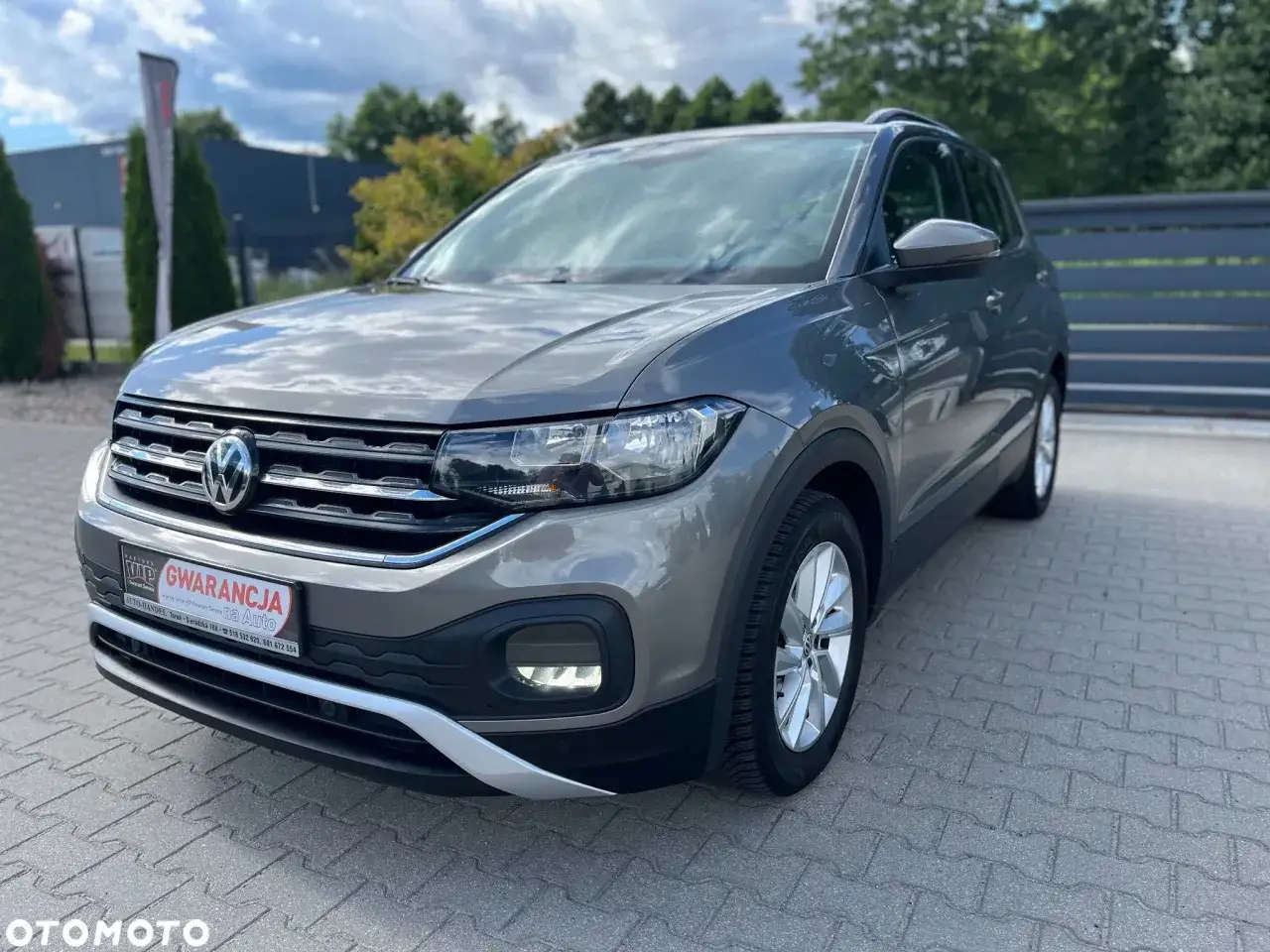 Szary Volkswagen T-Cross z gwarancją, zaparkowany na kostce brukowej przed budynkiem.