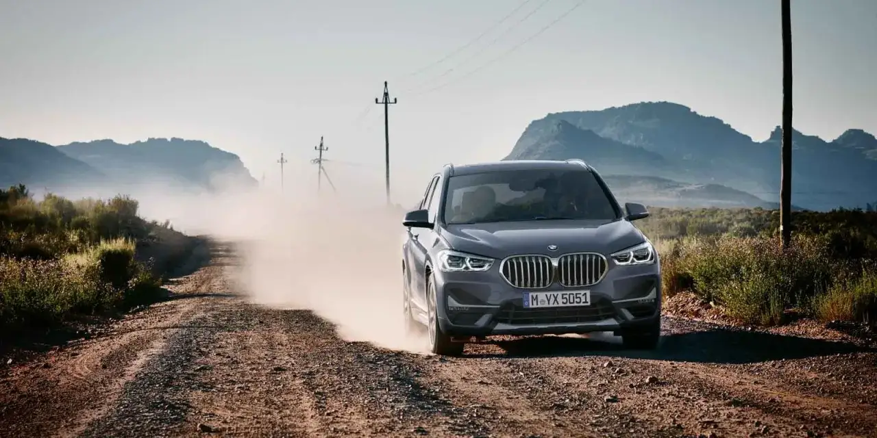 Szary BMW X1 2020 pędzi po piaszczystej drodze, wzbijając tumany kurzu. W tle góry i słupy energetyczne.