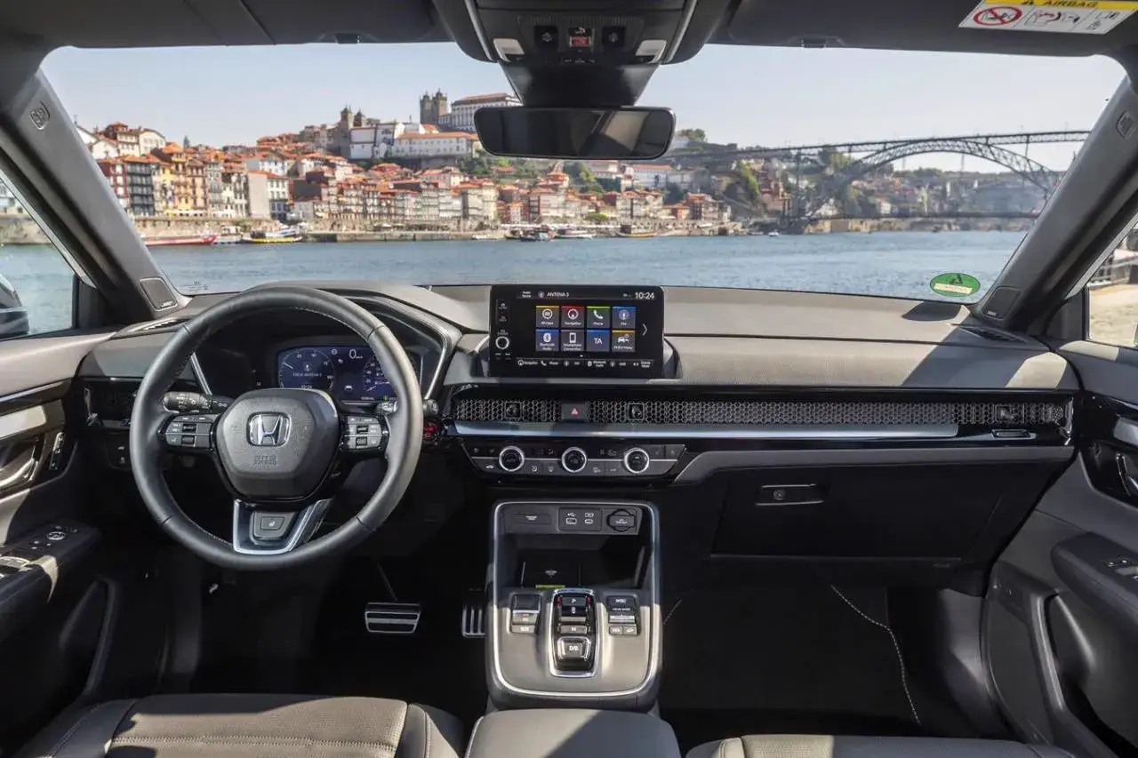 Wnętrze Hondy CR-V Hybrid z widokiem na malownicze miasto nad rzeką. Nowoczesna deska rozdzielcza i ekran multimedialny.