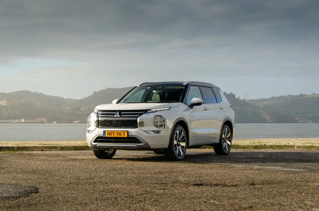 Biały Mitsubishi Outlander PHEV stoi nad wodą, z pagórkami w tle.