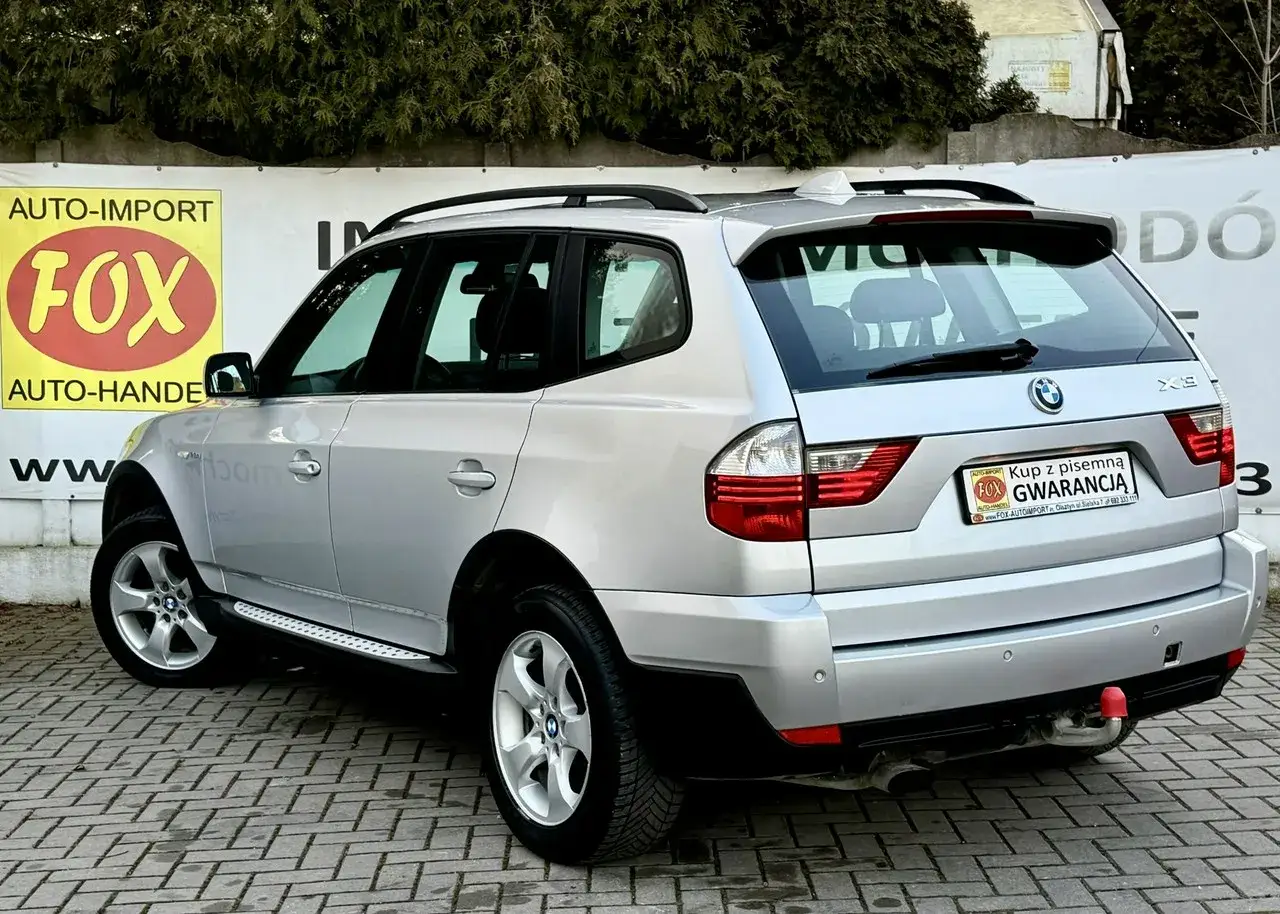 Srebrne BMW E83 z napędem na cztery koła, zaparkowane na kostce brukowej. Z tyłu widoczny hak holowniczy i tablica z gwarancją.