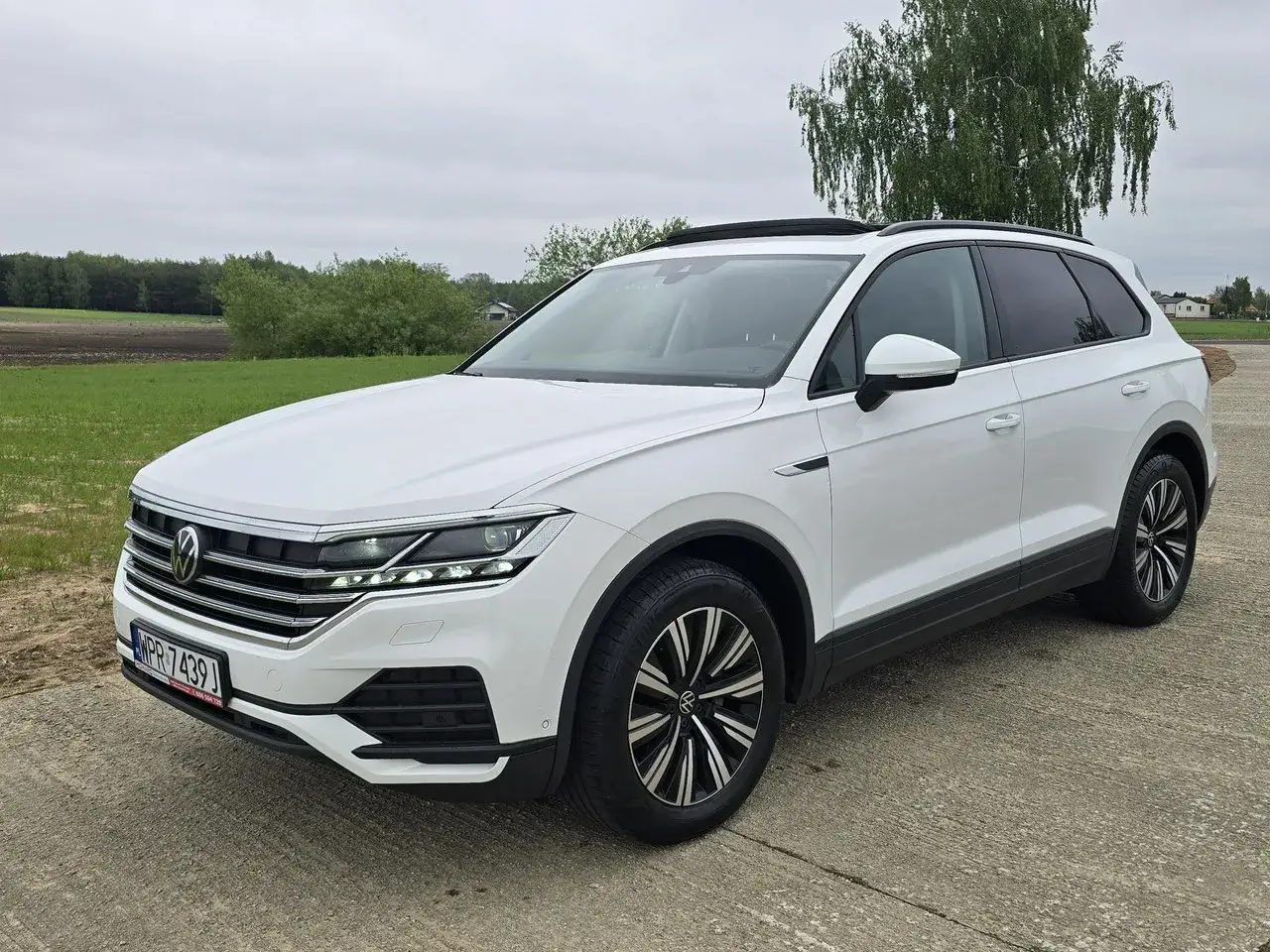Biały Volkswagen Touareg 3.0 TDI na tle zielonych pól i drzew. Elegancki SUV gotowy na każdą podróż.