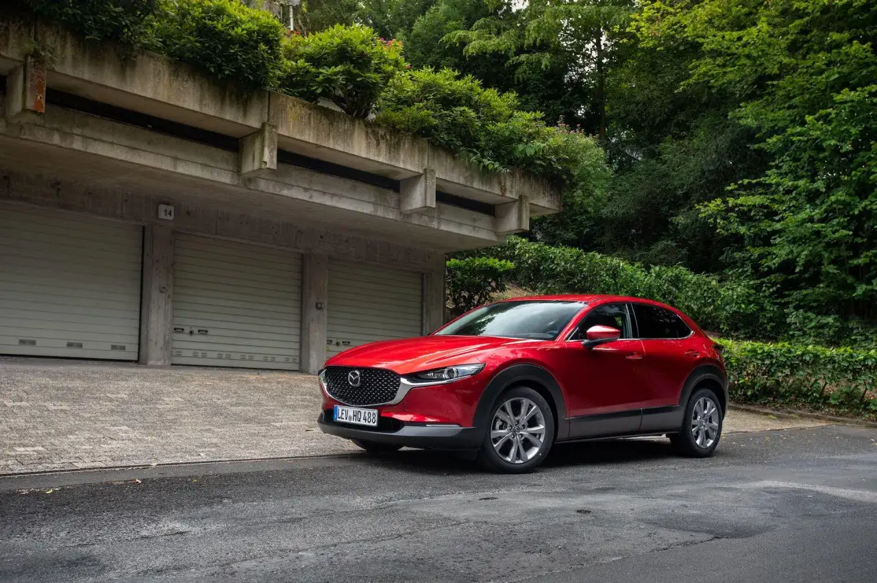 Czerwona Mazda CX-30 zaparkowana przed betonowymi garażami, otoczona bujną zielenią.