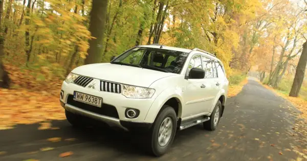 Biały model terenówki Mitsubishi Pajero Sport jedzie po drodze wśród jesiennych drzew.