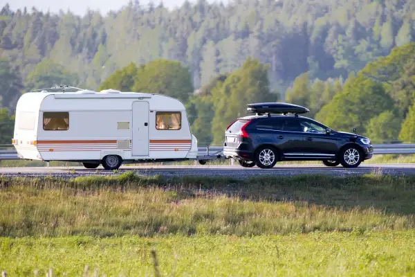 Ciemny SUV ciągnie białą przyczepę kempingową po drodze, na tle lasu.